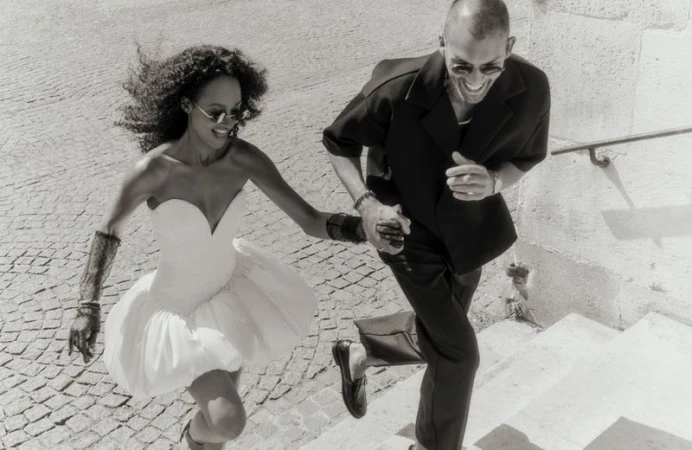 A fashionable couple is running up stone steps outside, with the woman in a short white dress and the man in a black suit.