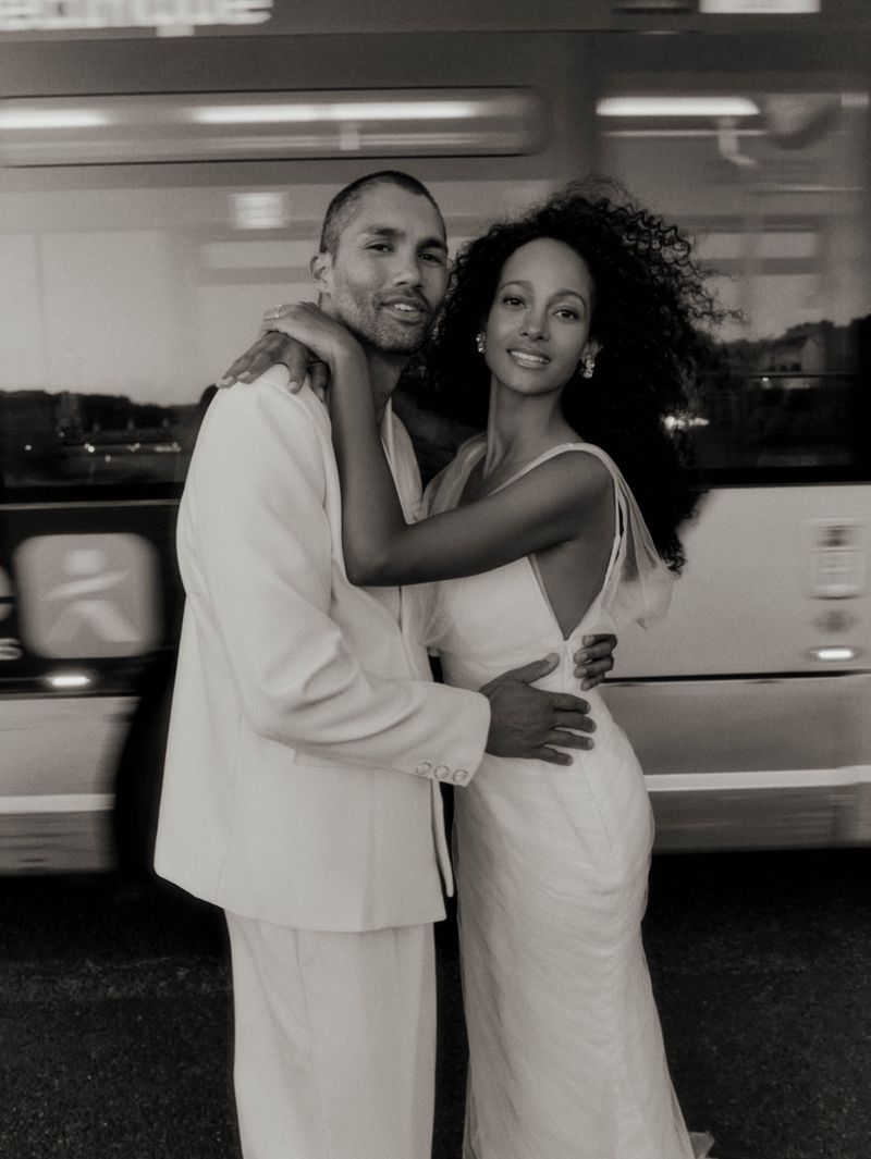 Monochrome portrait of a man in a white suit and a woman in a white dress embracing in front of a blurred bus