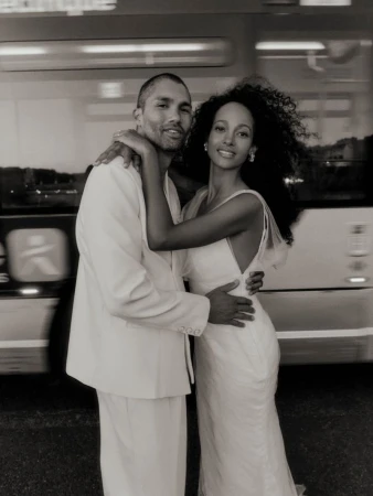 Monochrome portrait of a man in a white suit and a woman in a white dress embracing in front of a blurred bus