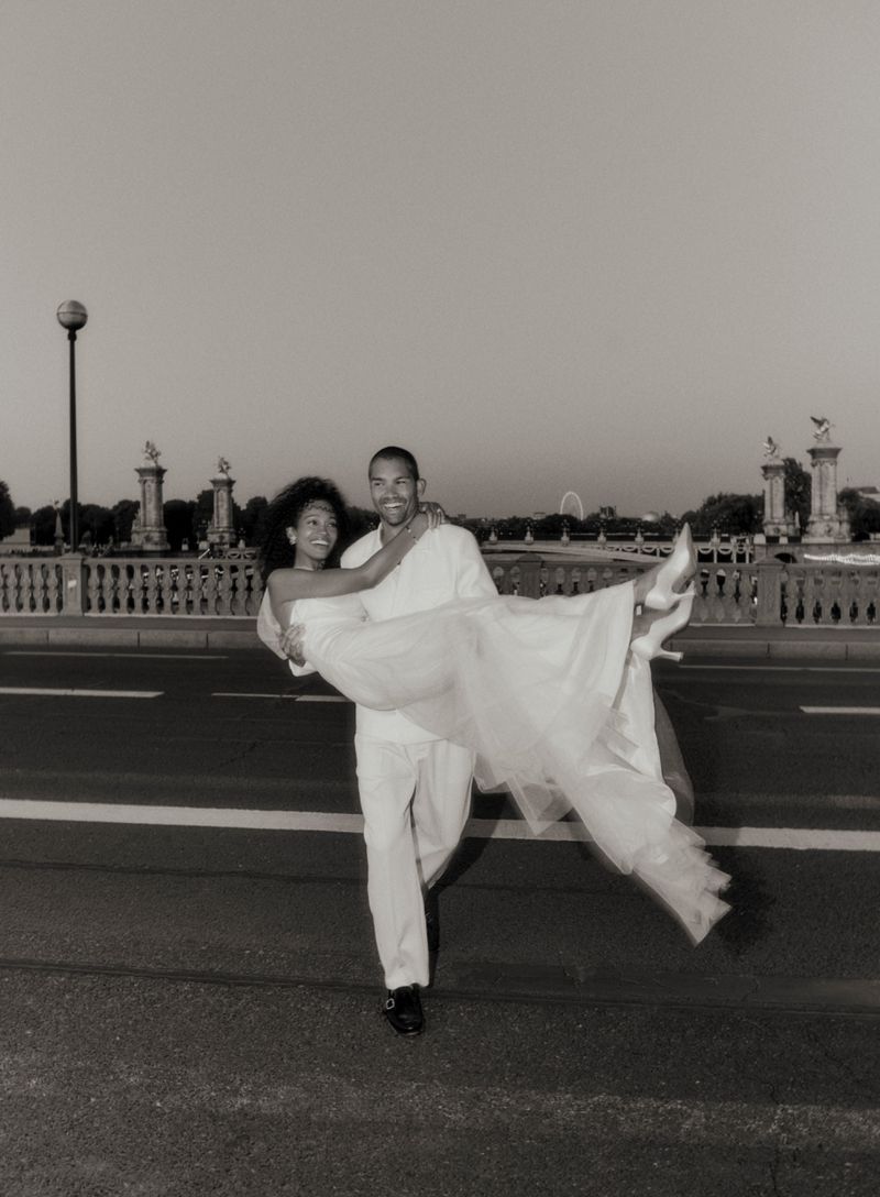 A man carries a woman in his arms in front of an ornate bridge with decorative elements. The image has a vintage, black-and-white tone.