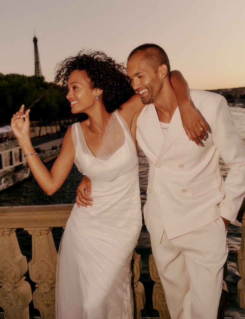Romantic couple poses in front of the Eiffel Tower in Paris at sunset. The woman wears a white dress; the man wears a white suit.