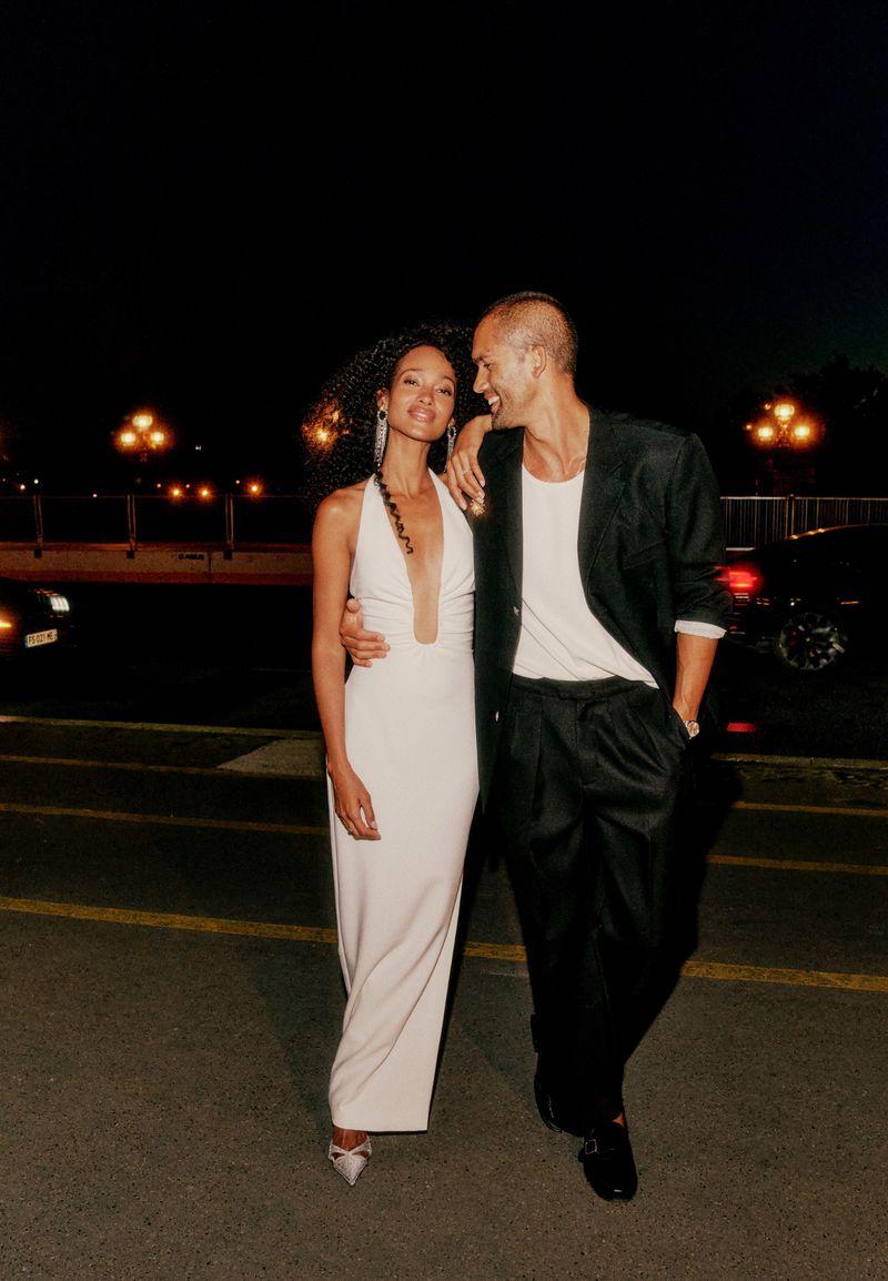 A fashionable couple poses at night. The woman wears a white halter dress and the man sports a blazer with loose pants and dress shoes.
