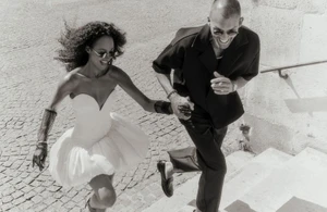 Black and white image of a smiling woman in a white dress and man in a dark shirt running down cobblestone outdoor steps.