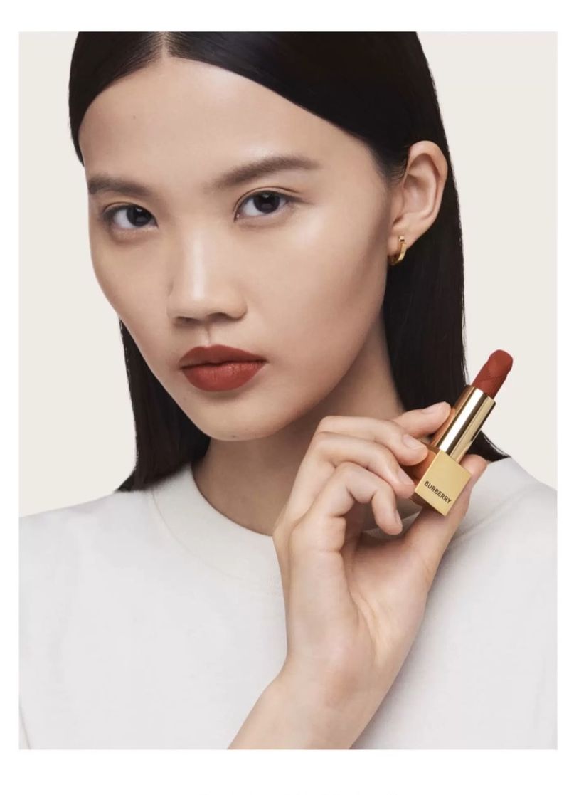 Asian woman with brown lipstick holding gold tube of Burberry lipstick in studio with white background. Beauty, fashion, cosmetics.