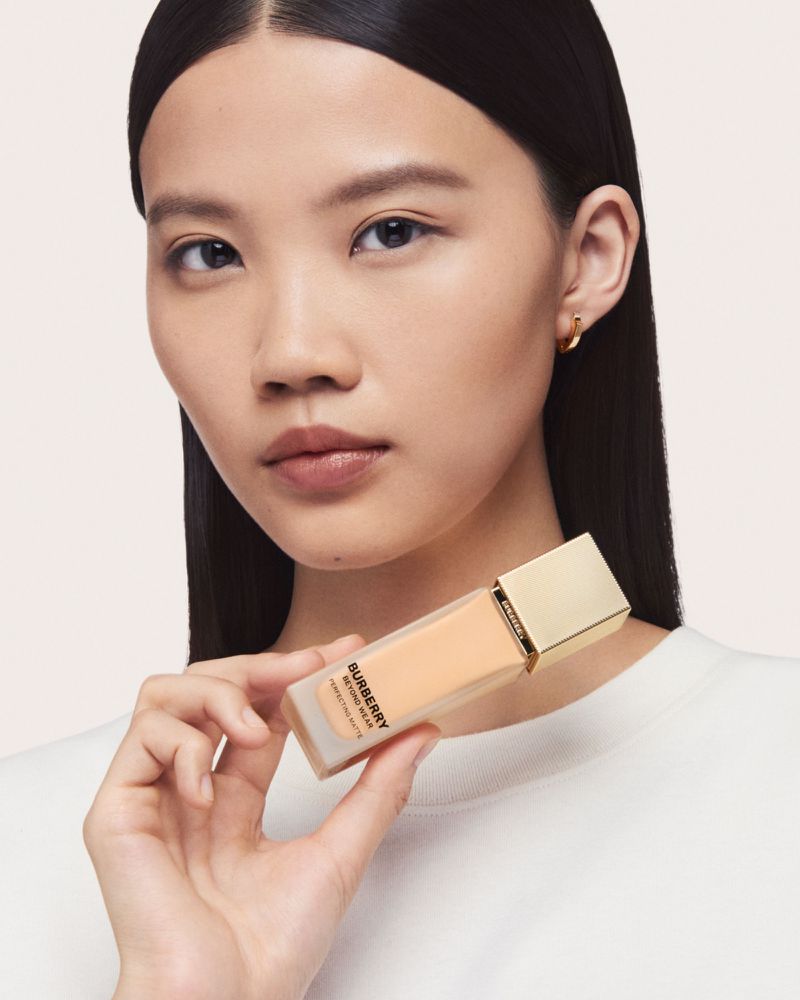 Asian woman holds Burberry Beyond Radiance foundation. Close-up studio shot of beauty product in fair complexion and light neutral background.
