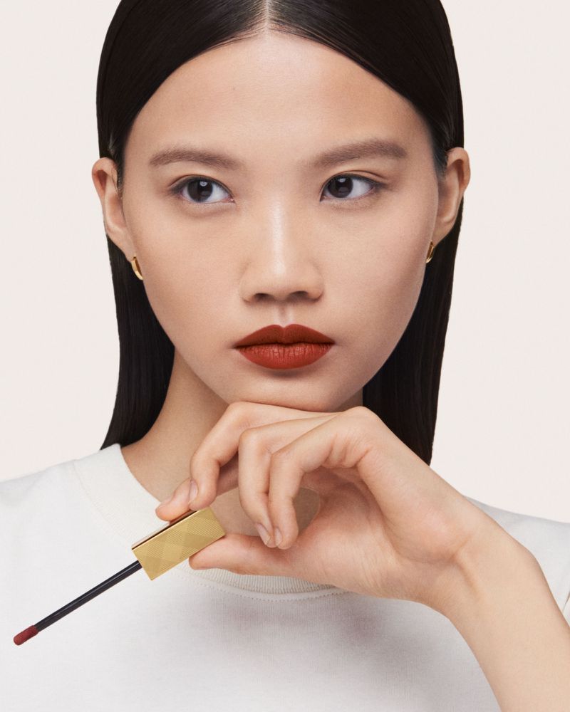 Asian woman showcases a red lipstick shade, holding the applicator, in a beauty-focused studio shot against a plain neutral backdrop.