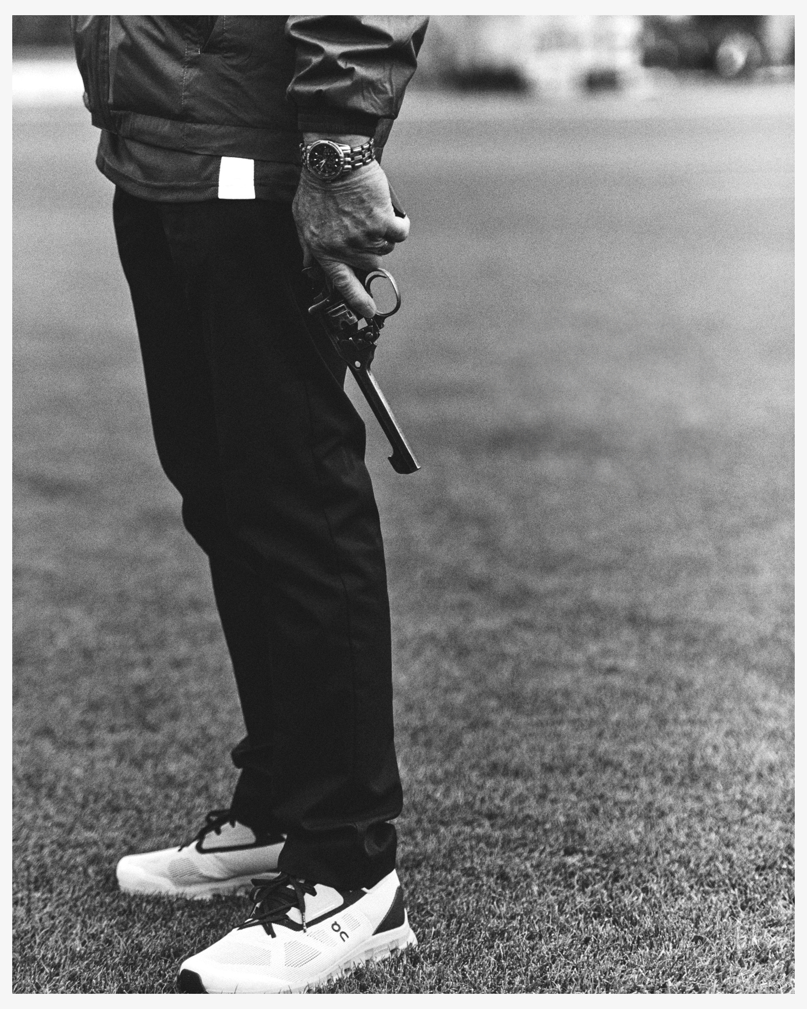 Black and white, medium shot of a person holding a starting pistol. The person is wearing athletic shoes, dark pants, and a jacket.