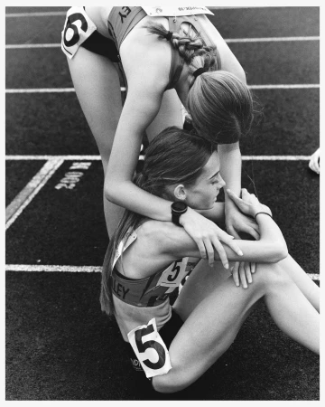 Two female athletes embrace on a running track. One athlete sits on the track while the other stands behind her with arms around her.