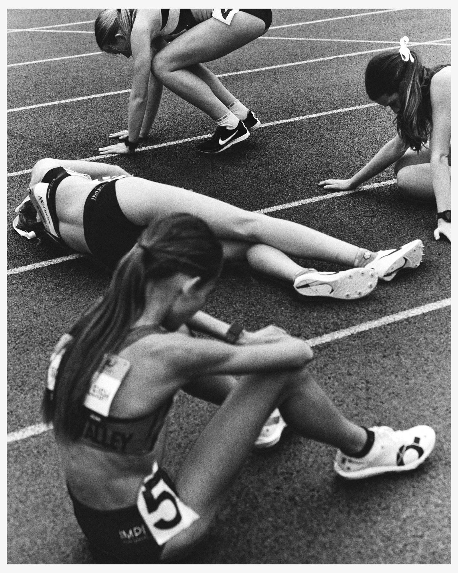 Black and white photograph shows four female athletes resting on a running track after a race, displaying exhaustion and athletic wear.