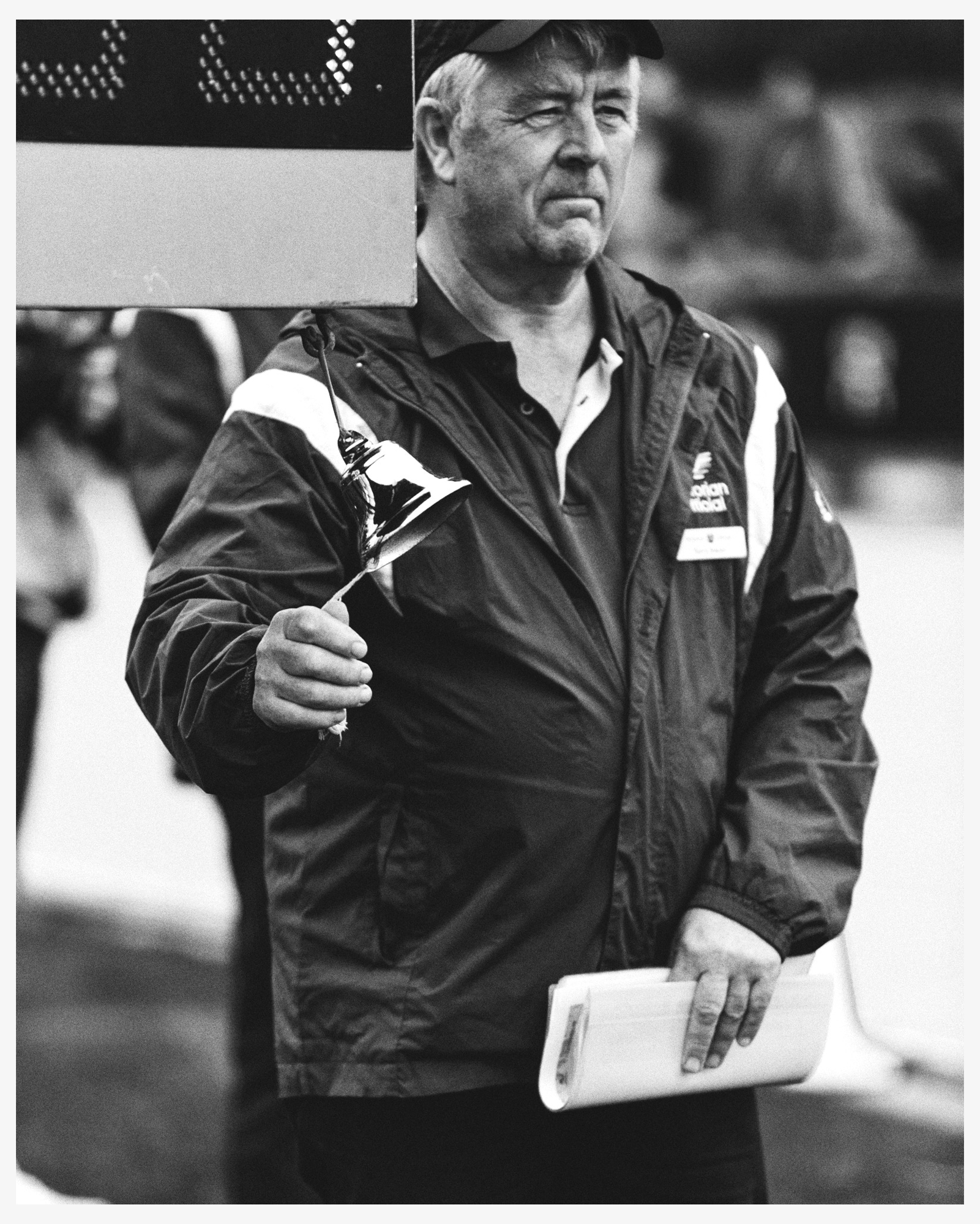 Black and white shot of a coach in jacket and cap holding a trophy and newspapers on a cloudy day near an outdoor sports field.