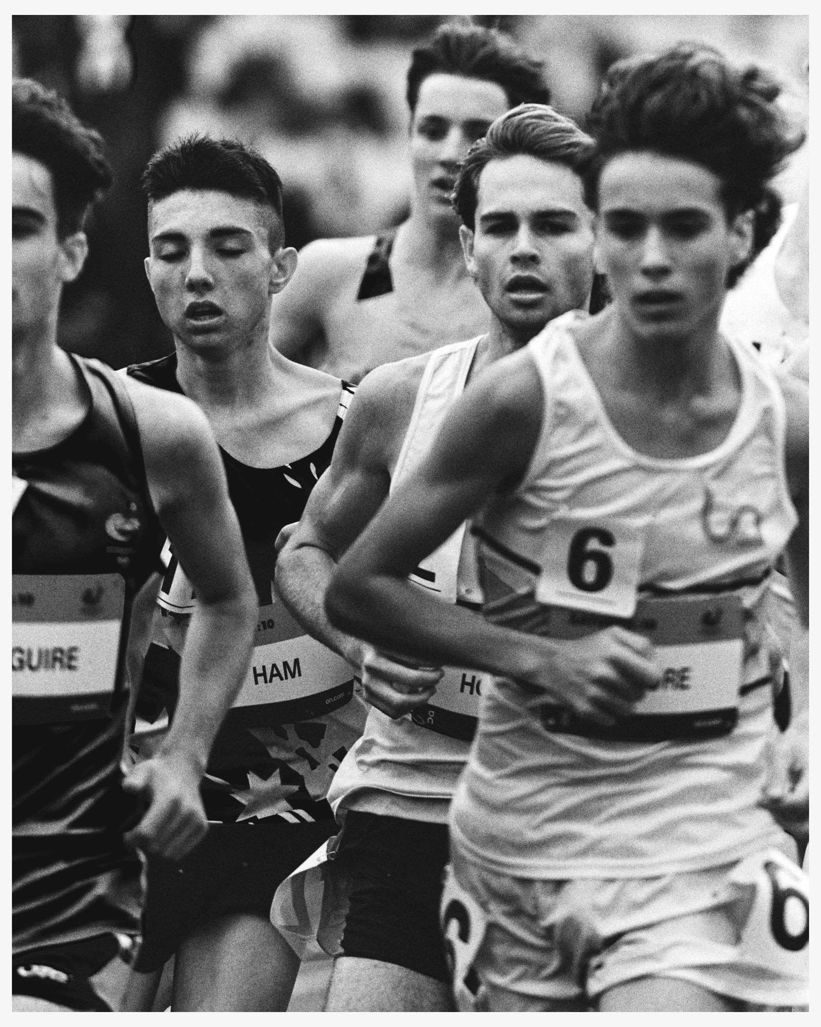 Black and white action shot of a group of runners competing in a race. The runners wear numbered bibs and athletic attire, captured outdoors.