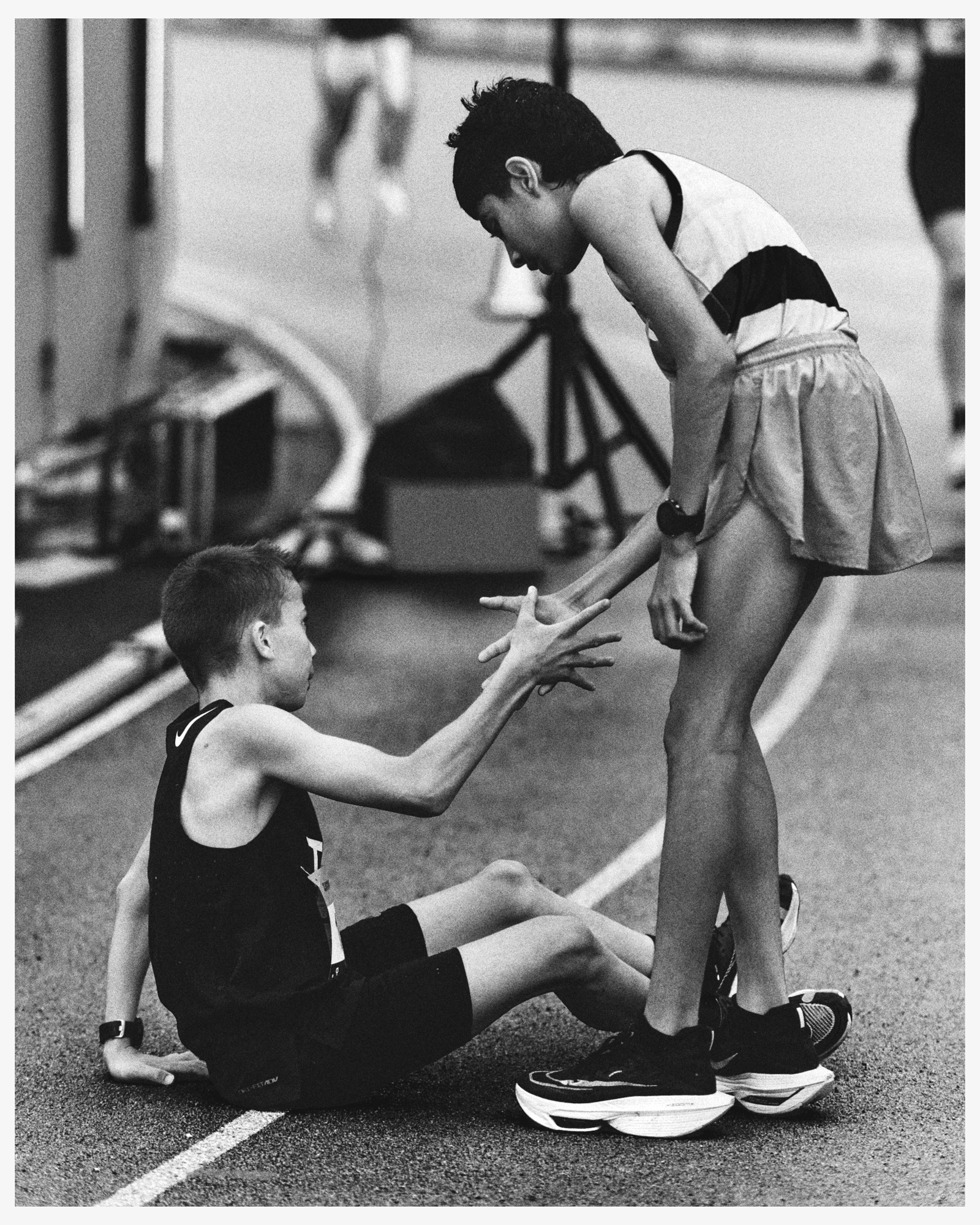 Black and white photo of two athletes in sportswear on a track, one sitting and reaching up to the other who is standing and offering a hand.