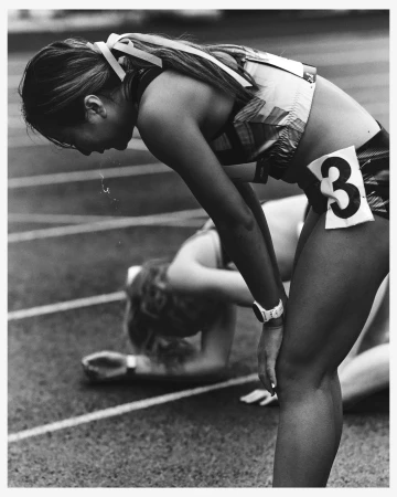 Black and white photo of a female athlete bent over with exhaustion, wearing a race number, with another athlete in the background.