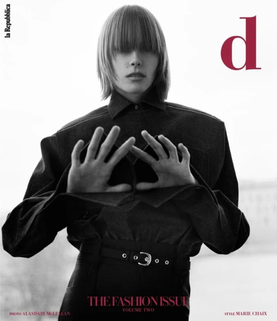 Editorial fashion shot features a person with blunt bangs, wearing a dark denim jacket and matching pants against a bright, diffused background.
