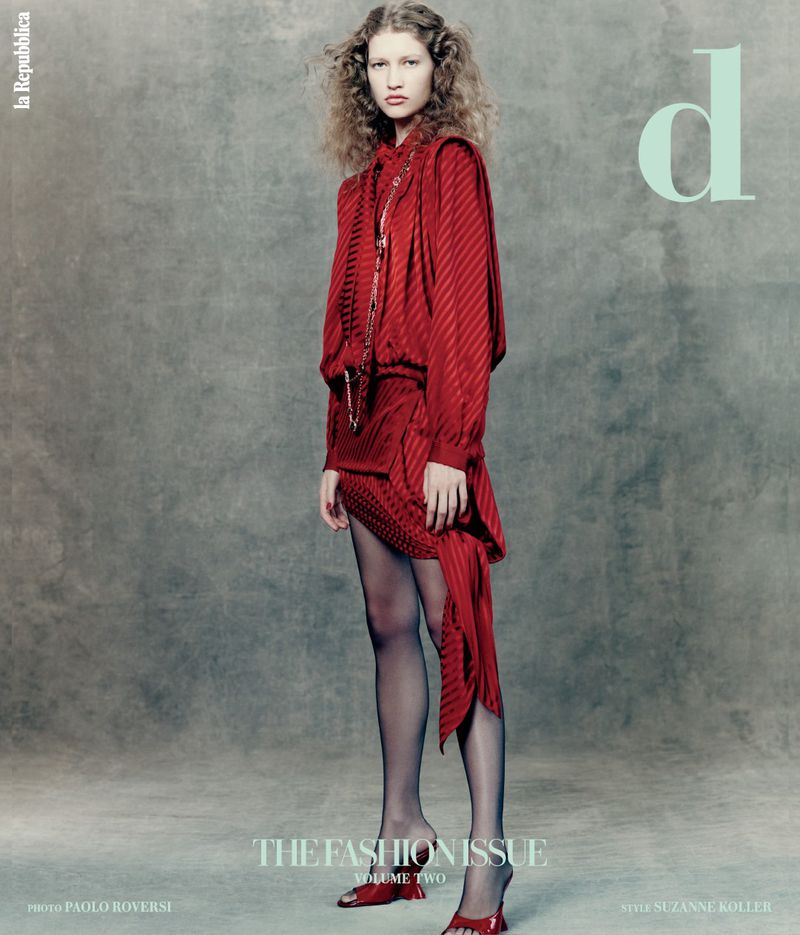 Fashion editorial: a female model with curly hair poses in a red, patterned outfit with black tights against a neutral grey background.