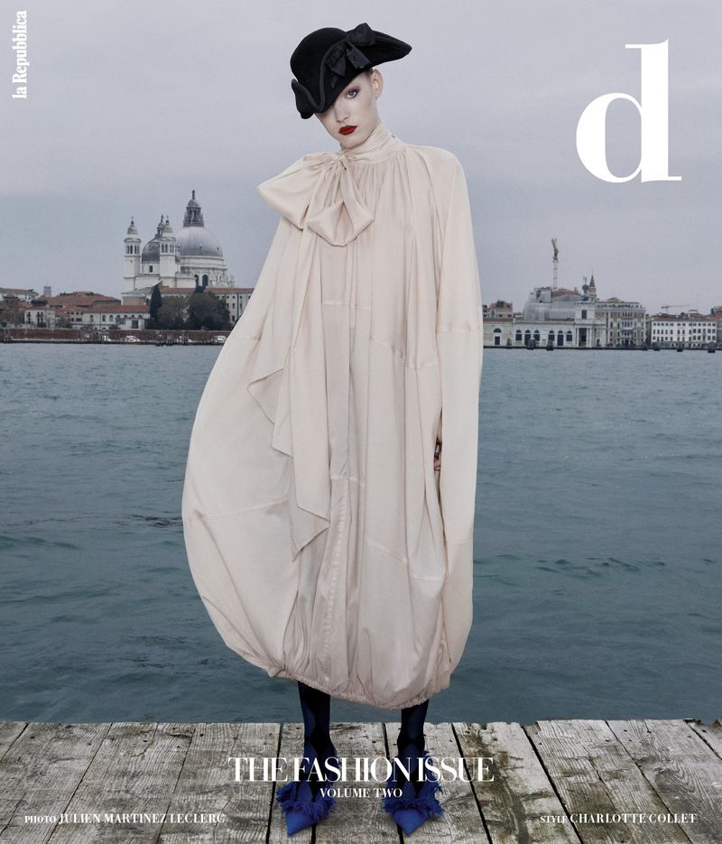 Model standing on a wooden dock wearing an oversized cream dress and black hat against a water and city background