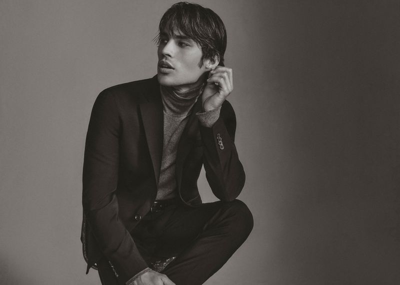 A young adult male model with dark hair poses in a grey turtleneck sweater and black suit in a studio shot against a grey background.