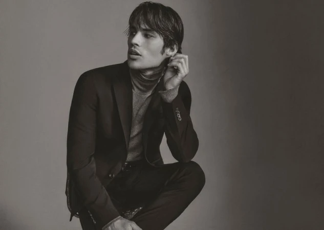 A young adult male model with dark hair poses in a grey turtleneck sweater and black suit in a studio shot against a grey background.