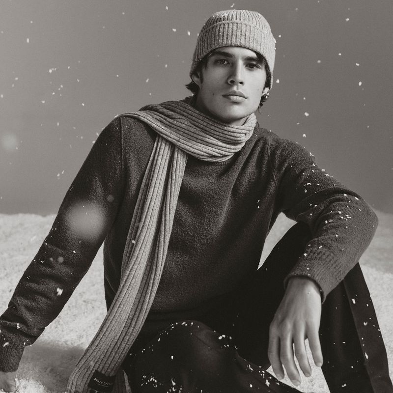 Monochrome portrait of a young adult male wearing a beanie, sweater, and scarf sitting in a winter scene with snow falling.
