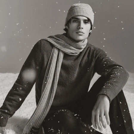 Monochrome portrait of a young adult male wearing a beanie, sweater, and scarf sitting in a winter scene with snow falling.