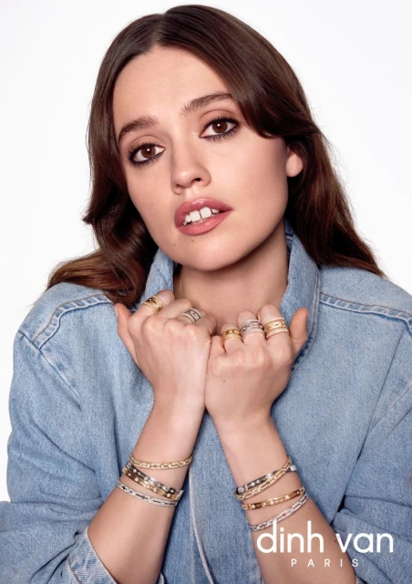 Close-up shot of a model wearing a denim jacket and displaying jewelry on her fingers and wrists against a white backdrop.