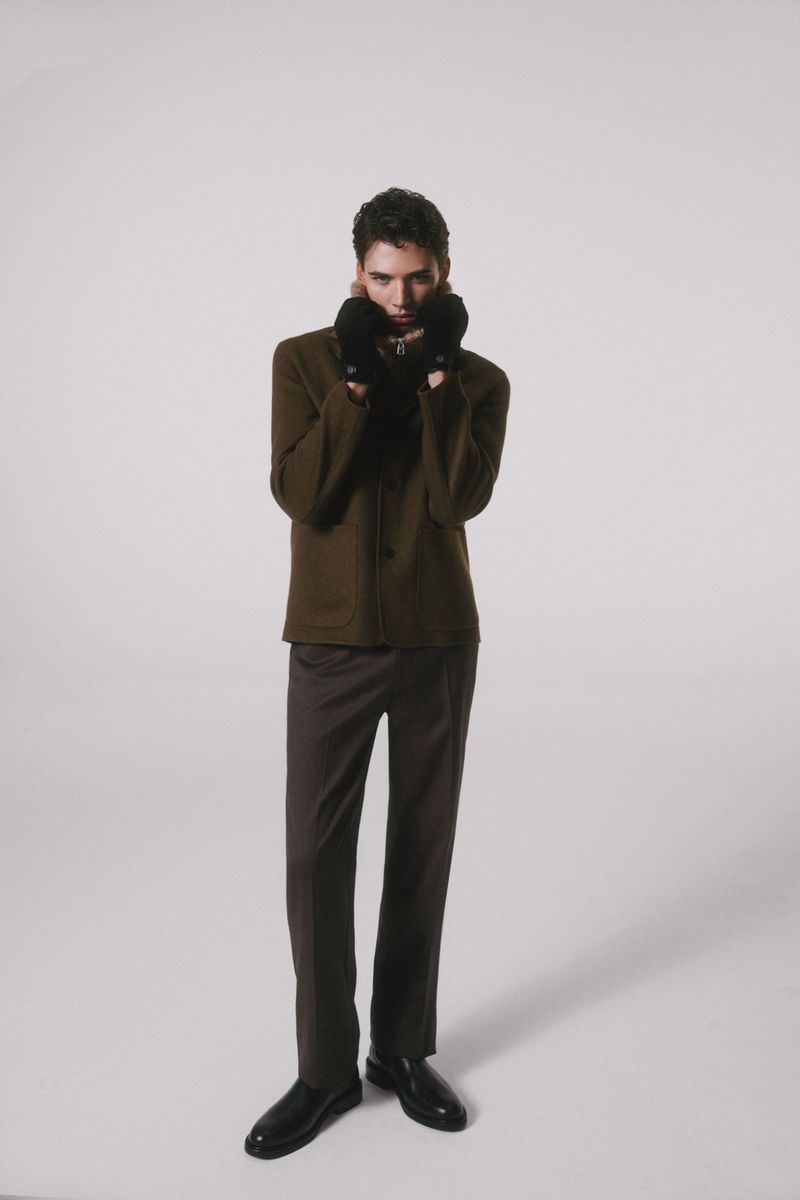 A full-length portrait of a young man indoors, wearing an olive jacket with brown trousers and black shoes, fashionably posing in soft lighting.