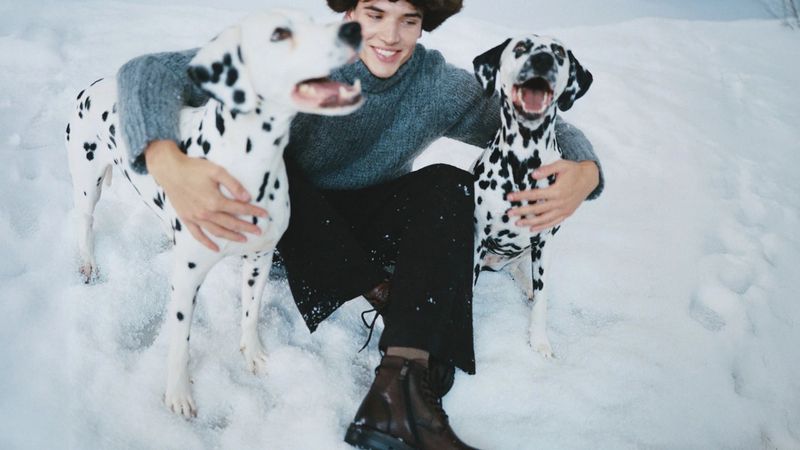 Young man smiles while seated in the snow, embraced by two Dalmatian dogs, wearing a gray sweater, dark pants, and brown boots.