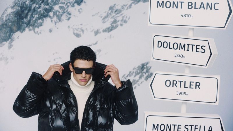 Man in black puffer jacket with sunglasses poses in front of a snowy mountain backdrop and signpost with mountain names.