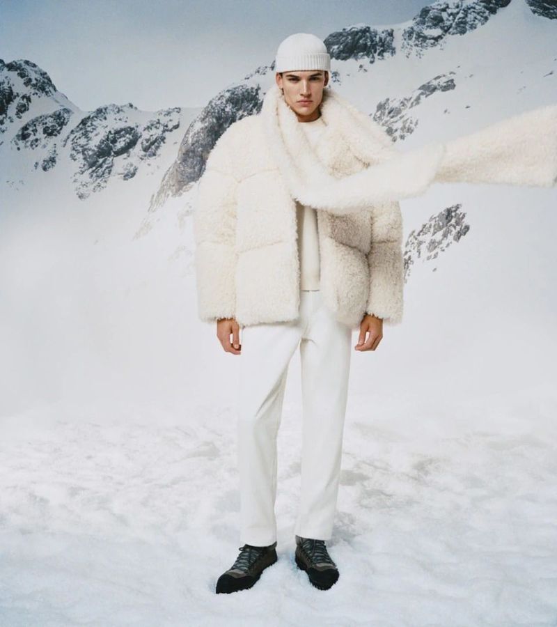 A fair-skinned adult male in a white knit hat, scarf, coat, sweater and trousers stands in a snowy mountain landscape.