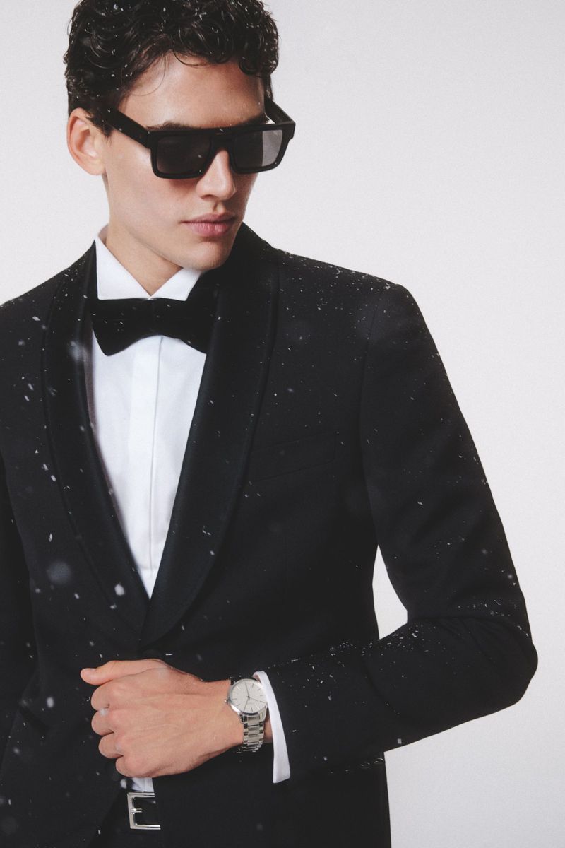 Portrait of a handsome adult male wearing a black tuxedo, bow tie, silver watch, and black sunglasses against a white backdrop.