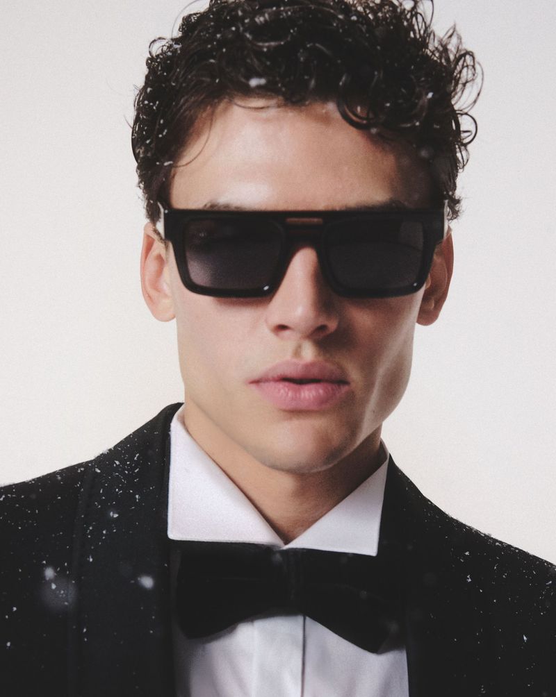 Close-up of a stylish adult male wearing sunglasses, a white shirt, and a black bow tie, accessorized with a black tuxedo jacket.