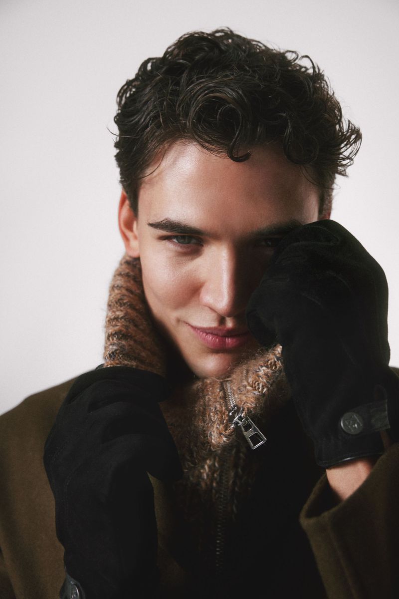 Close-up portrait of a young adult man with curly dark hair wearing a brown wool coat, gloves, and a turtleneck in a studio setting.