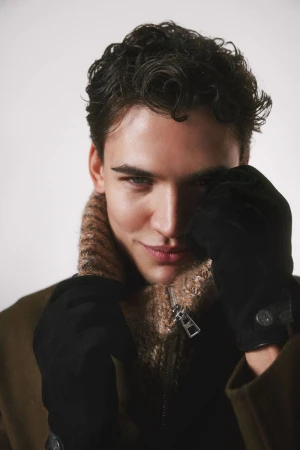 Close-up portrait of a young adult man with curly dark hair wearing a brown wool coat, gloves, and a turtleneck in a studio setting.