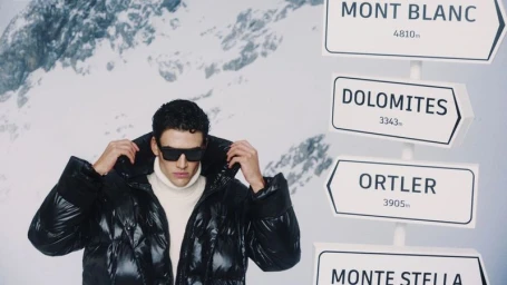 Model posing in a black glossy puffer jacket, white turtleneck, and sunglasses next to mountain trail signposts.