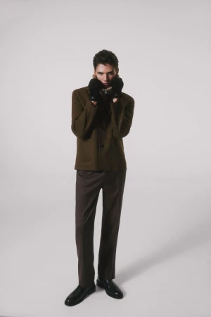 Tomasz Labis poses in an olive brown jacket, dark trousers, black gloves, and polished black shoes against a light grey studio backdrop.