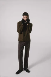 Tomasz Labis poses in an olive brown jacket, dark trousers, black gloves, and polished black shoes against a light grey studio backdrop.