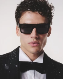 Model Tomasz Labis wearing black rectangular sunglasses and a black tuxedo with bow tie against a white background with falling snow.