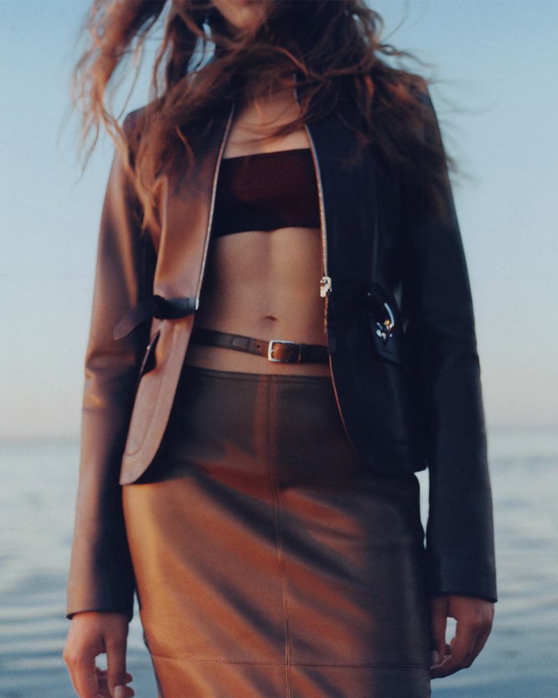 Woman poses in black jacket and brown skirt near calm water. Natural light with soft focus on hair and horizon under a serene sky.
