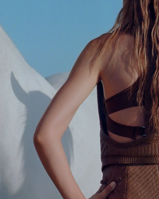 Back view of a woman in a brown quilted vest and skirt against a light blue background with white geometric shapes.