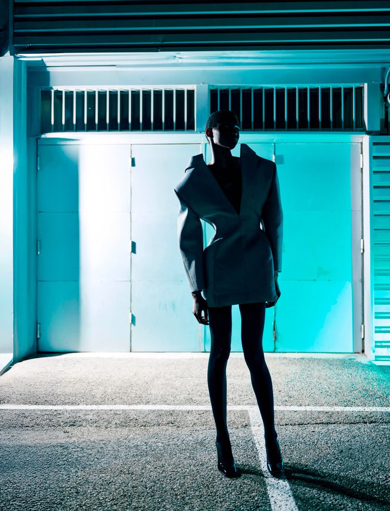 Woman in a grey jacket and black tights stands in front of a building with closed doors under bluish lighting, fashion editorial.