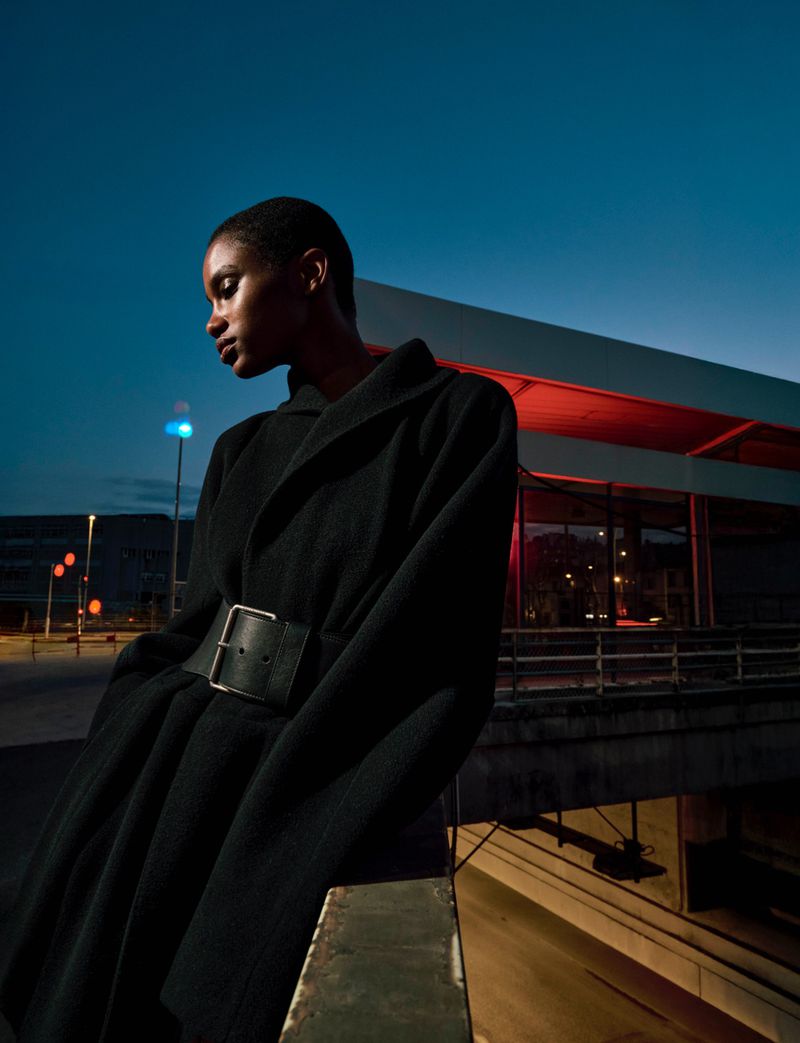 A low angle shot of a black woman with short hair wearing a black belted coat, standing against an urban backdrop with an illuminated building.