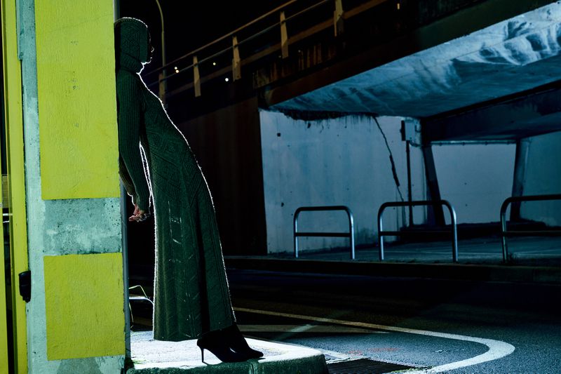 A woman in a long hooded sweater dress stands against a wall in an urban setting at night, illuminated by yellow and blue lights.