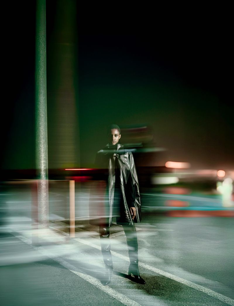 Fashion image of a woman in a black coat standing on a crosswalk at night with motion blur and dark green and orange streetlights.