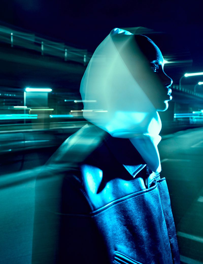 Profile view of a Black woman in a futuristic hooded coat, with light streaks and blurred city lights in the background in a fashion editorial.