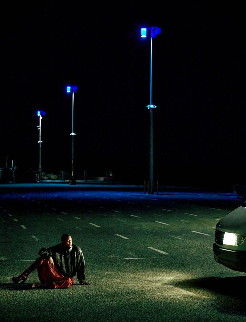 Person sitting on asphalt at night near vehicle and blue street lights, wearing a black sweatshirt and red skirt in an open space.