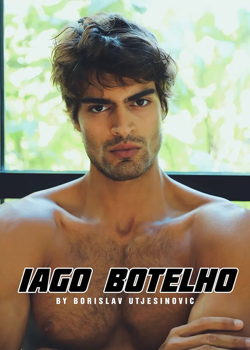 Shirtless adult male poses indoors near a window with soft, natural lighting. The man has brown hair, dark eyes, and a light beard.