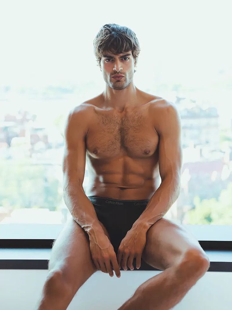 Shirtless muscular man sits against a bright window wearing Calvin Klein underwear in a studio shot with chest and abs visible.