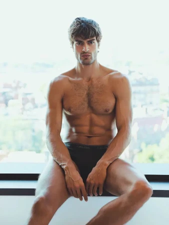 Shirtless muscular man sits against a bright window wearing Calvin Klein underwear in a studio shot with chest and abs visible.