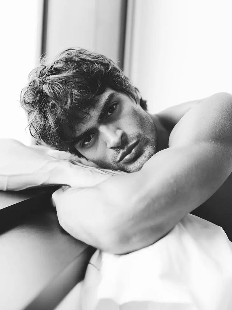 Black and white close-up of a handsome adult male resting his arms on a pillow looking directly at the camera with a neutral expression.