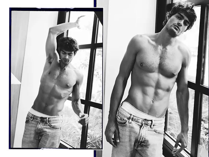 Black and white studio shot of a shirtless adult male model in jeans posing by a window, showcasing a fashion and editorial aesthetic.